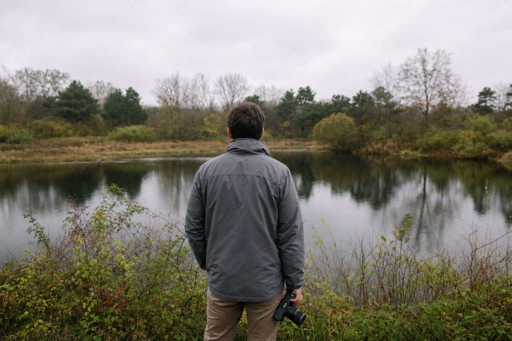 A person seen from behind at a low overlook above a quiet pond and tree line