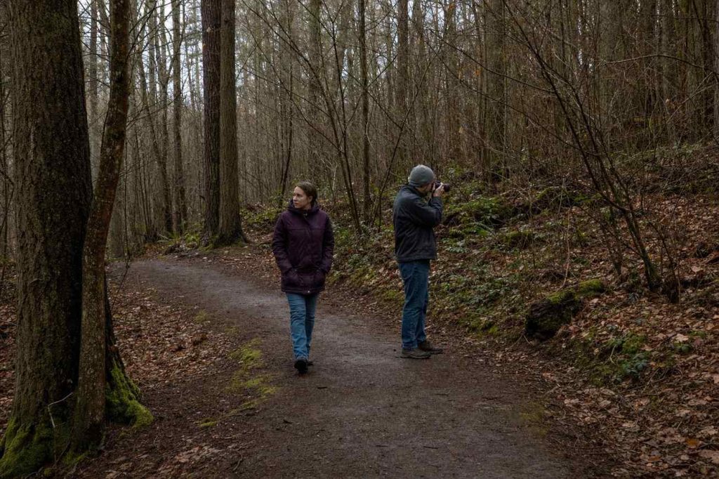 Two people walking slowly along a wooded path, one pausing to photograph