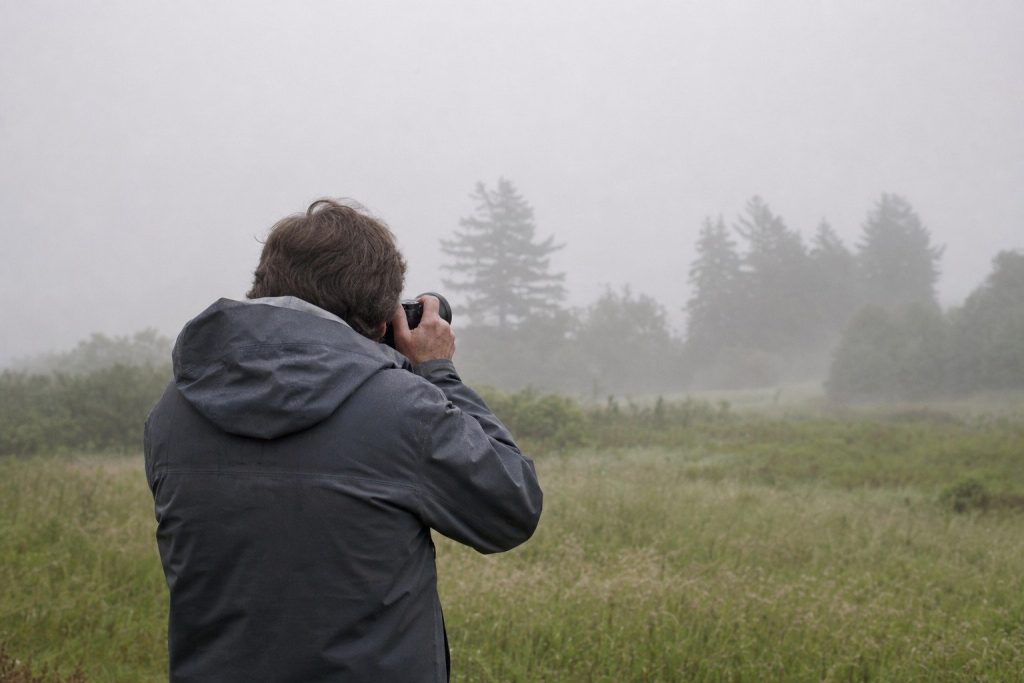 A photographer in a rain jacket seen from behind in a foggy field at dawn