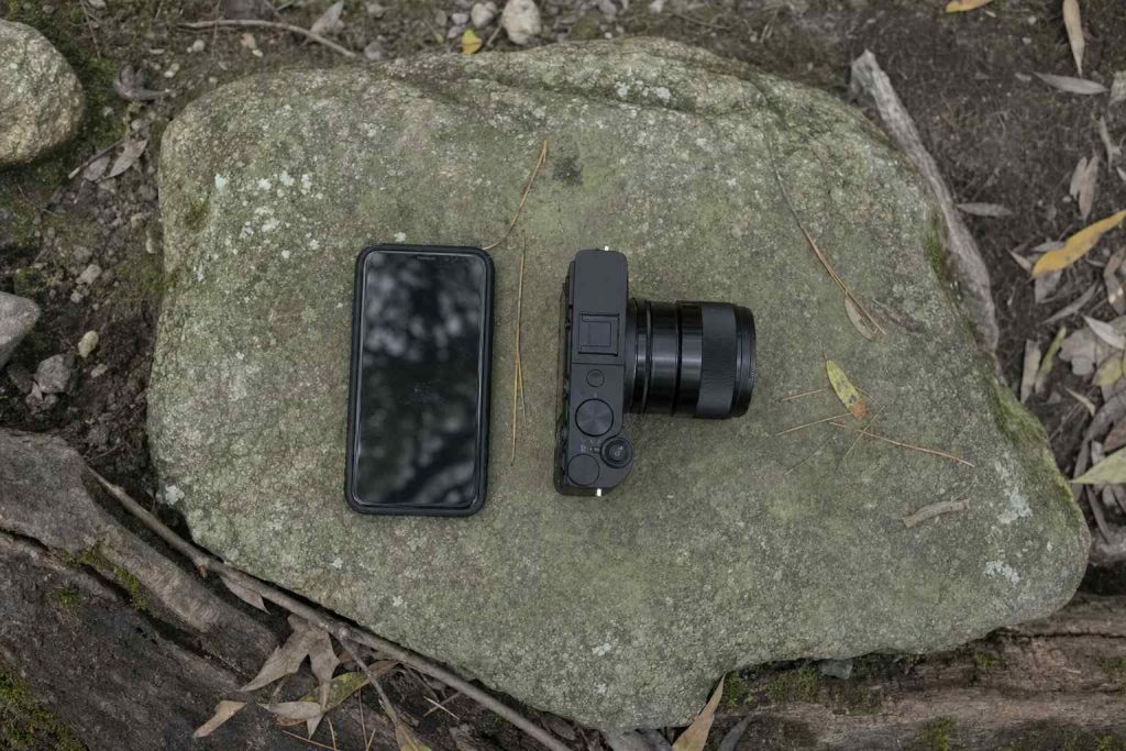 A smartphone and a small camera side by side on a mossy trail rock, shot from above